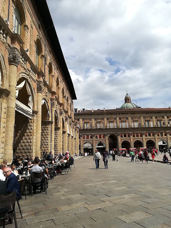 People enjoying an outdoor cafe at Palazzo d'Accursio, Piazza Maggiore, Bologna, under a cloudy sky.