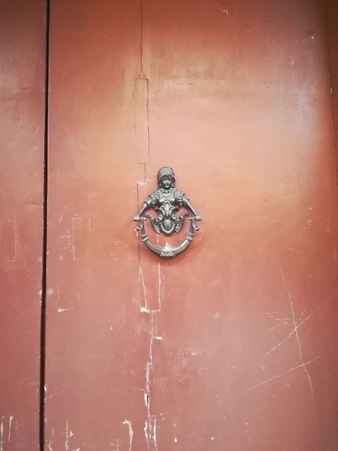 Ornate iron door knocker on weathered red door, featuring a detailed figure with classical design elements.