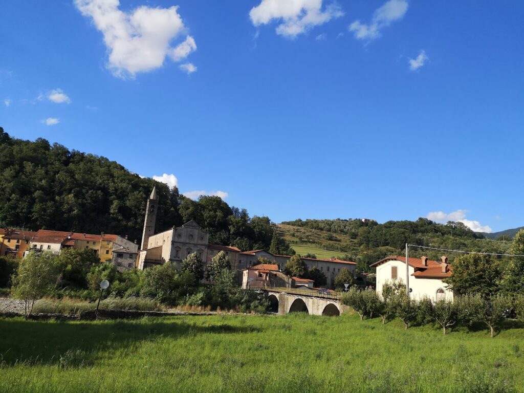Scenic Italian village with church, bridge, and green hills under a clear blue sky.