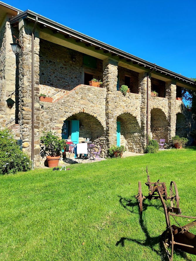 Rustic stone farmhouse with turquoise doors, sunny lawn, and vintage plow in Italy.