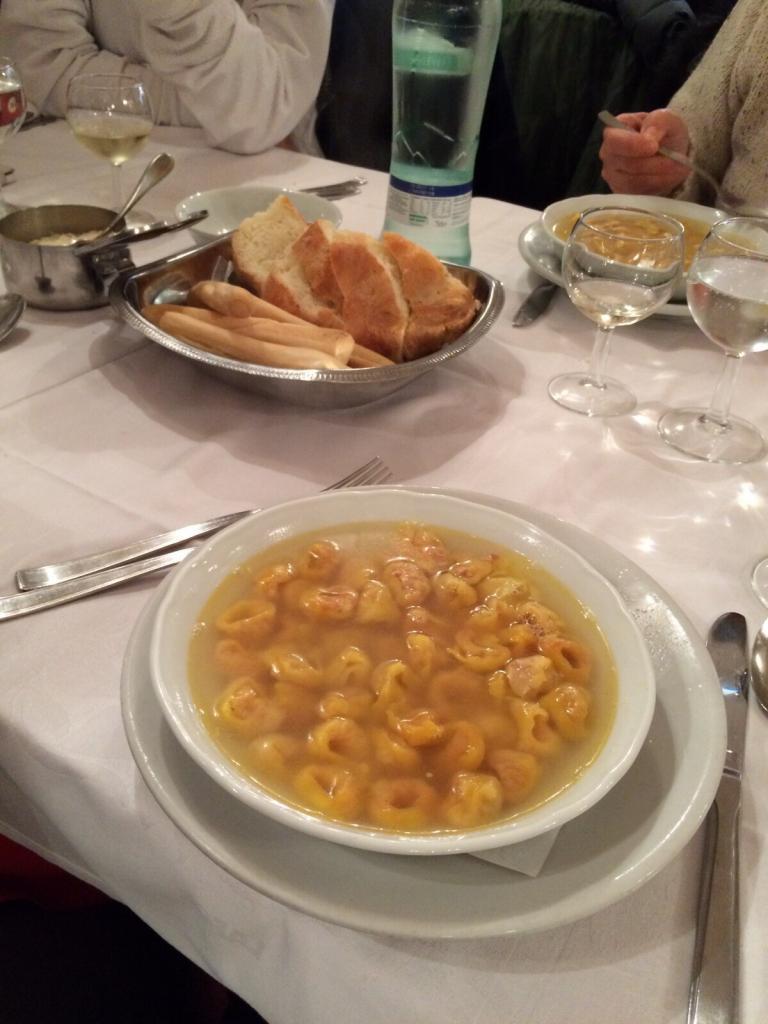 Bowl of tortellini soup and bread on a dining table with wine and sparkling water.
