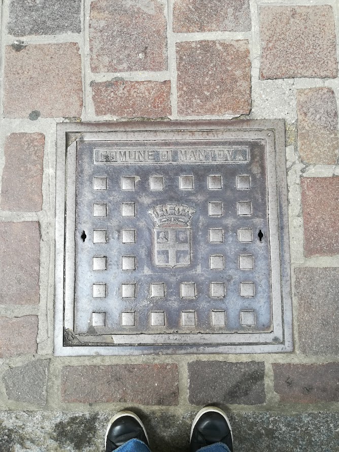 Manhole cover labeled Comune di Mantova with crest, surrounded by cobblestones, viewed from above.