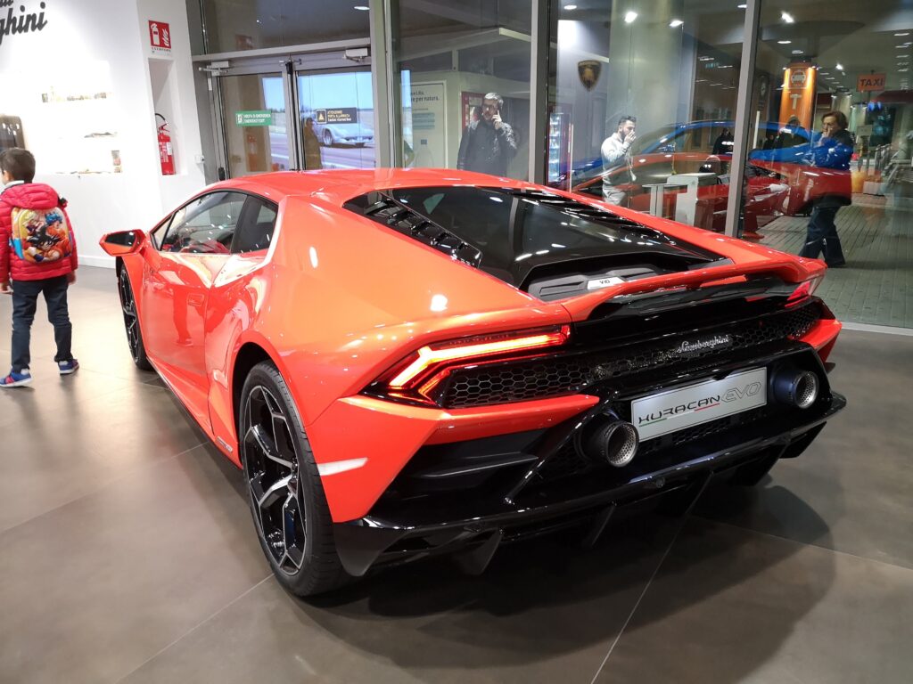 Bright orange Lamborghini Huracan EVO on display in a showroom with people observing from inside and outside.