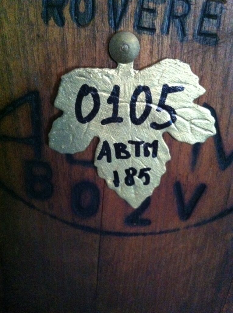 aceto balsamico tradizionale di Modena Golden leaf-shaped tag with 0105 ABTM 185 on a wooden barrel, background text partially visible.