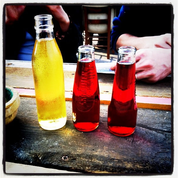 non alcohol aperitivo, three colorful glass bottles with beverages on a rustic table, with people sitting in the background.