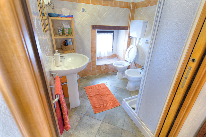 Compact bathroom with sink, toilet, bidet, shower, orange accents, and shelving next to a small window.