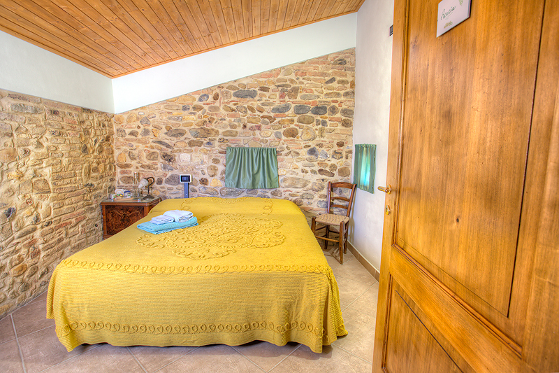 Cozy rustic bedroom with stone walls, wooden ceiling, yellow bedspread, and vintage chair.