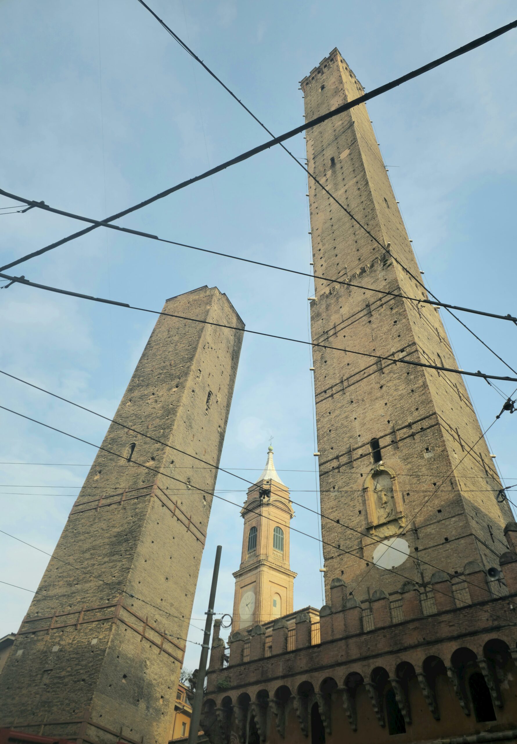 Two historic leaning towers in Bologna, Italy, against a clear blue sky with power lines crossing the view.