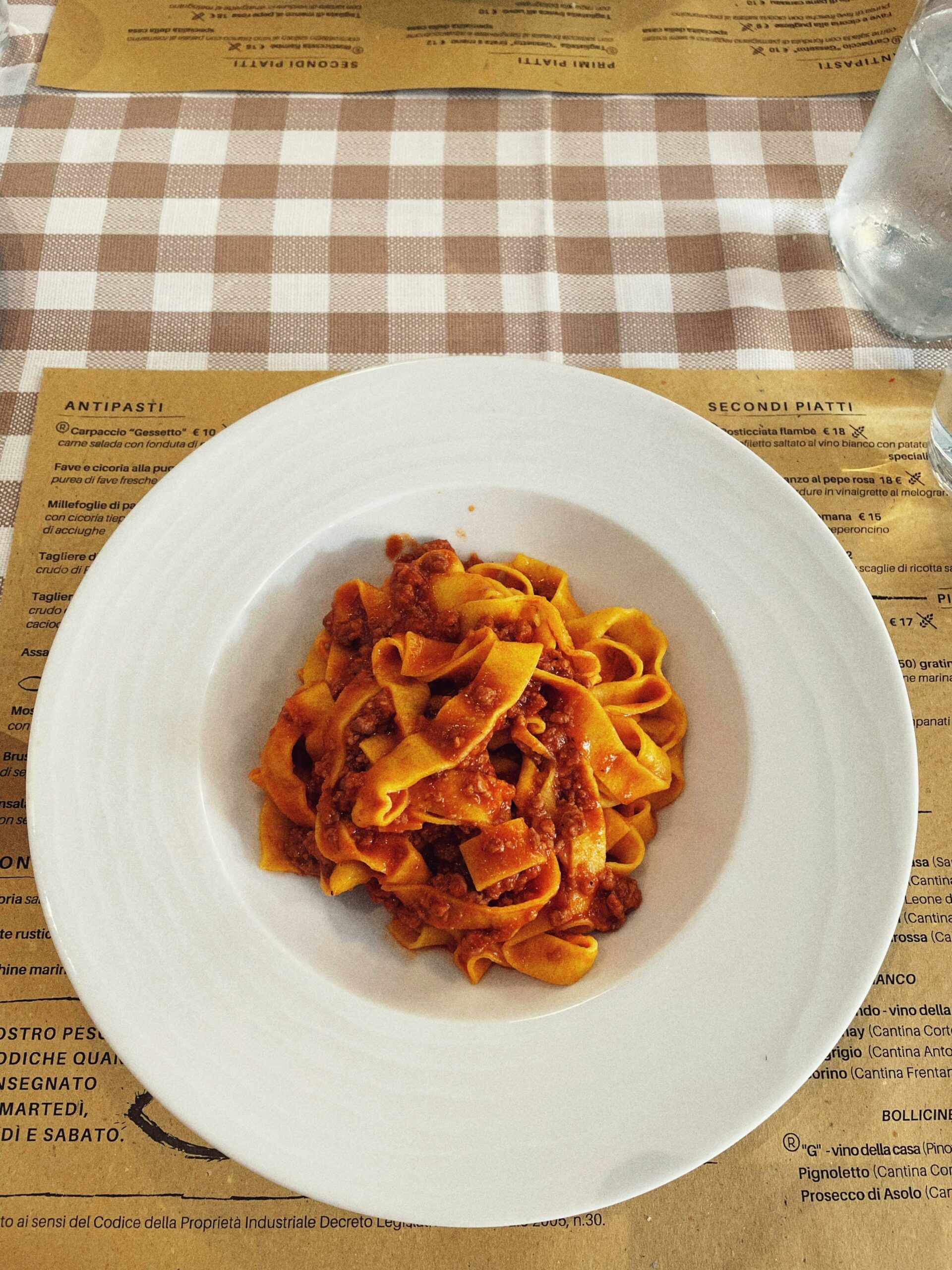 Plate of tagliatelle pasta with rich meat sauce on a brown checkered tablecloth in an Italian restaurant.