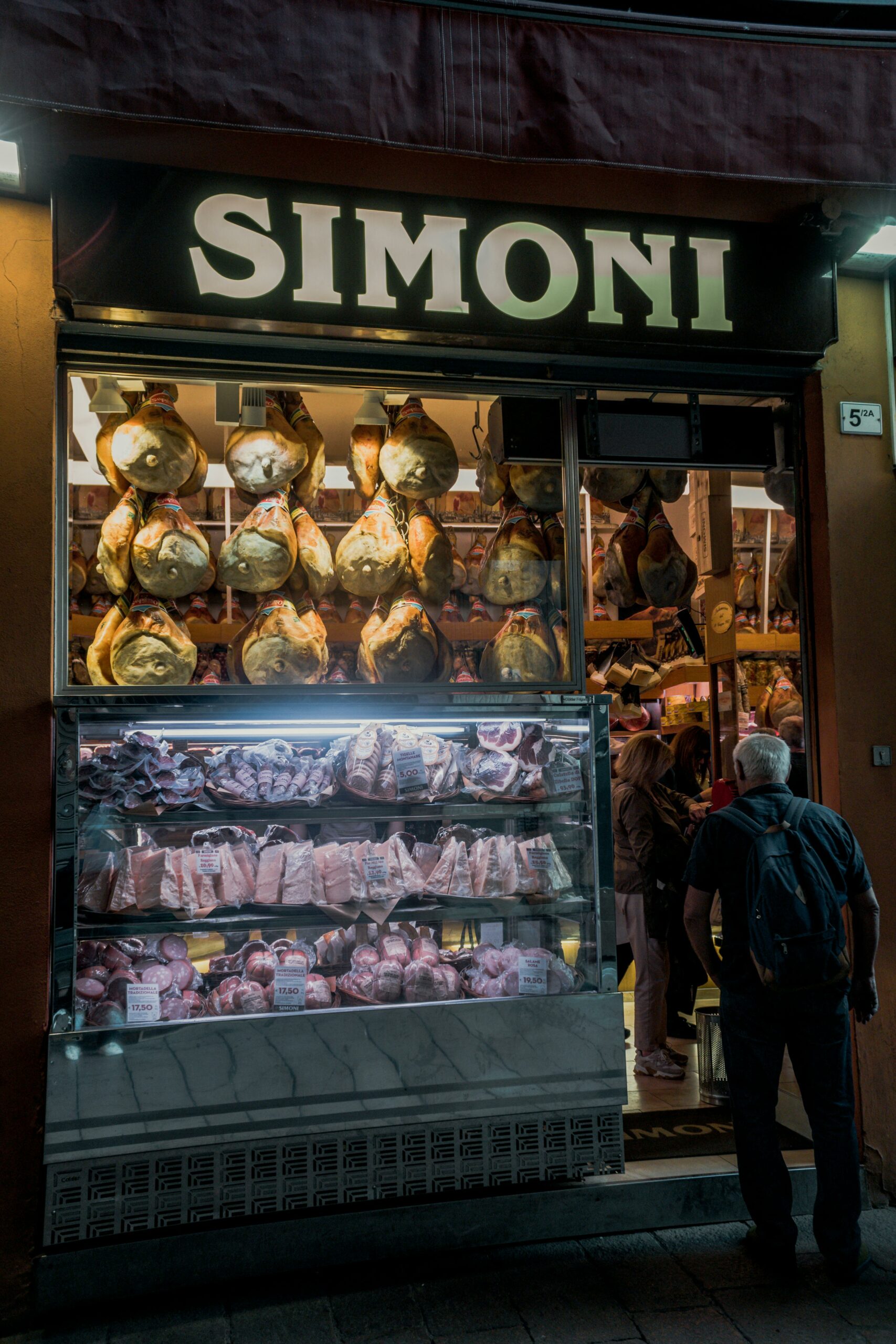 Deli shop with hanging cured meats and a selection of cuts in a window display, sign reads Simoni.