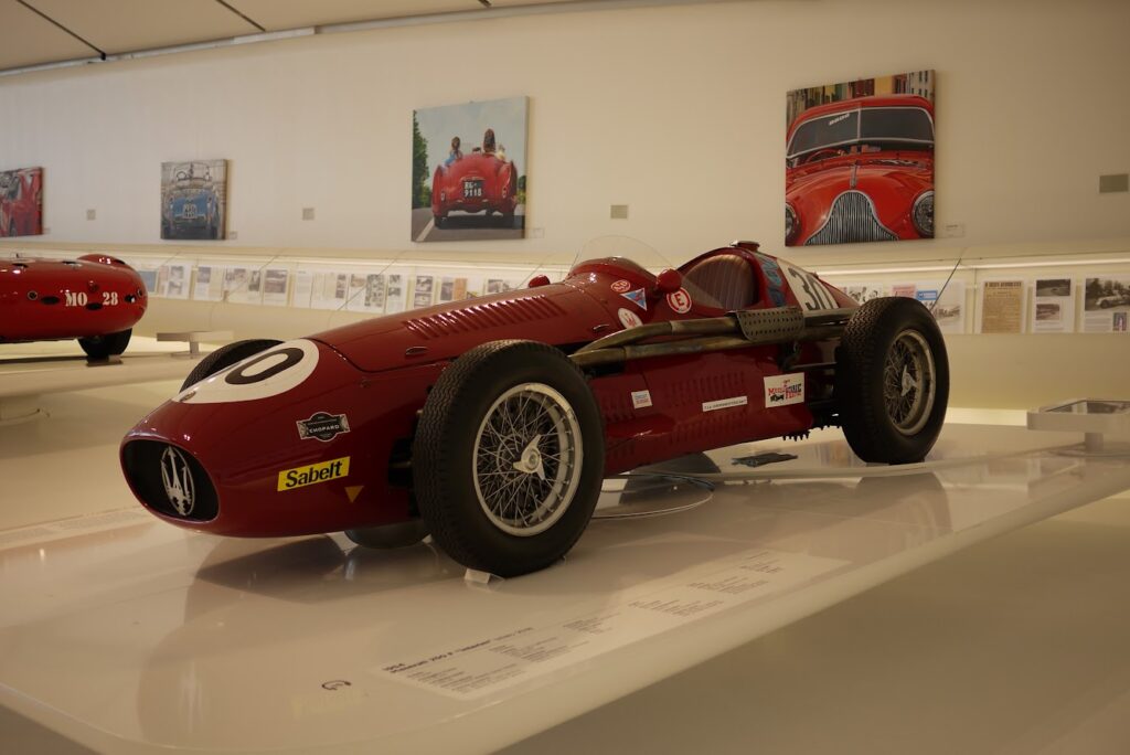 Red vintage Maserati race car in museum exhibit with art and historic displays in background.