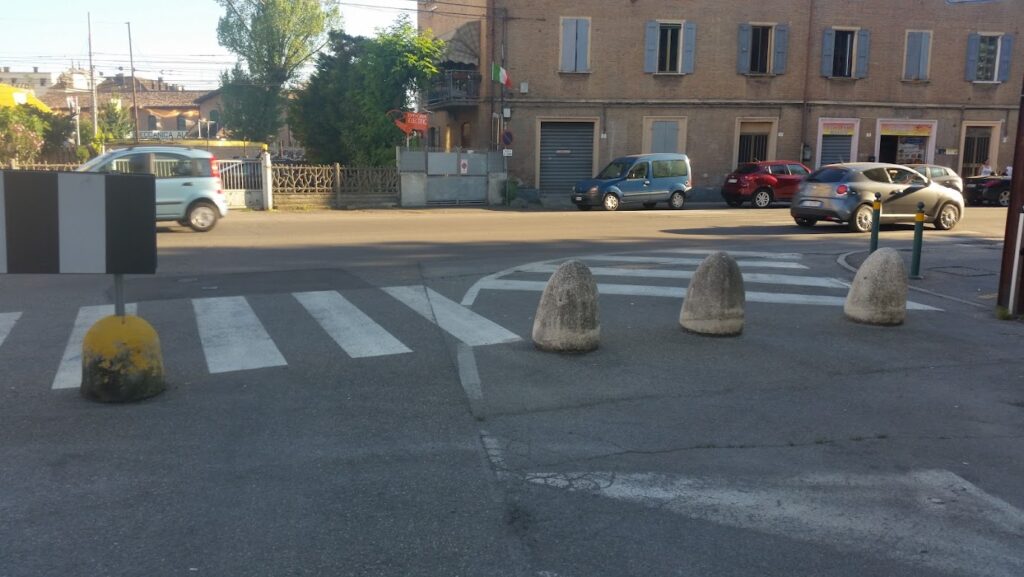 Street intersection with cars, crosswalk, and traffic barriers, set against a backdrop of buildings and trees.