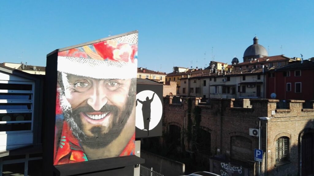 Colorful mural on a building with a smiling man's face, set against a backdrop of historic Modena architecture.
