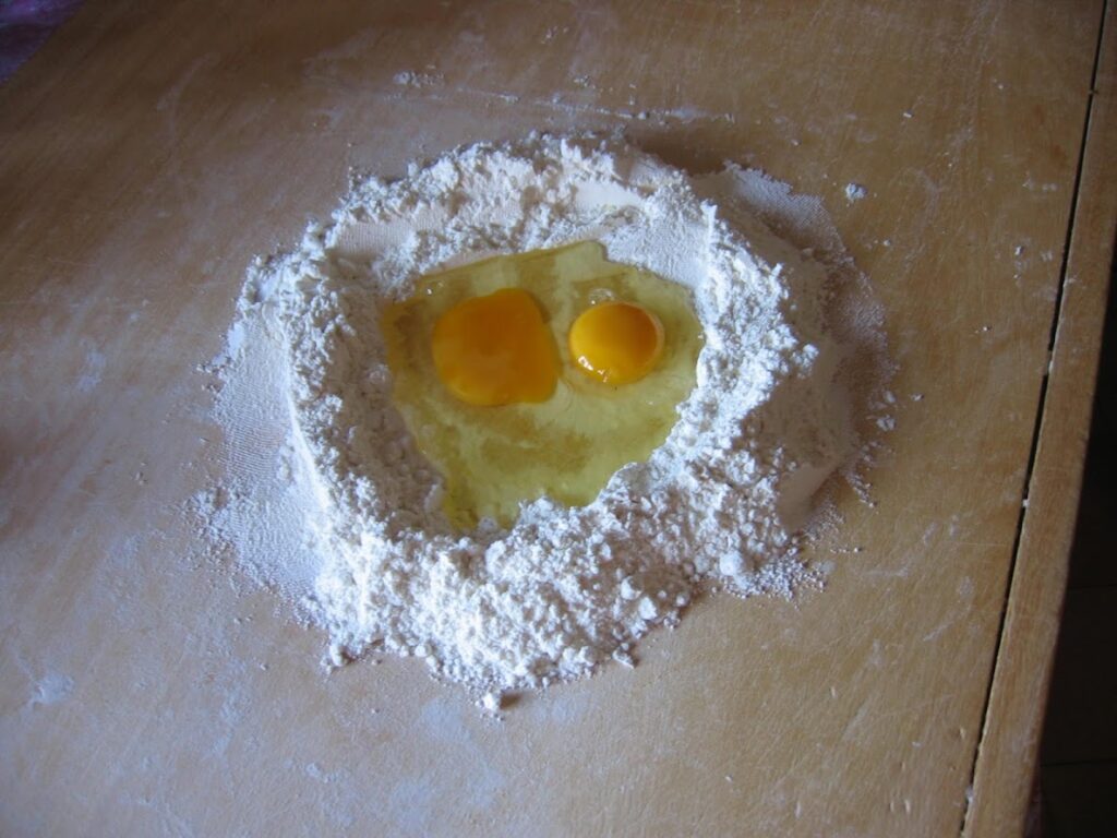 Flour mound with two cracked eggs on a wooden surface, ready for pasta dough preparation.
