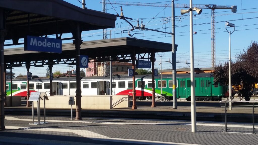 Train at Modena station platform under clear sky, with green and red train cars.
