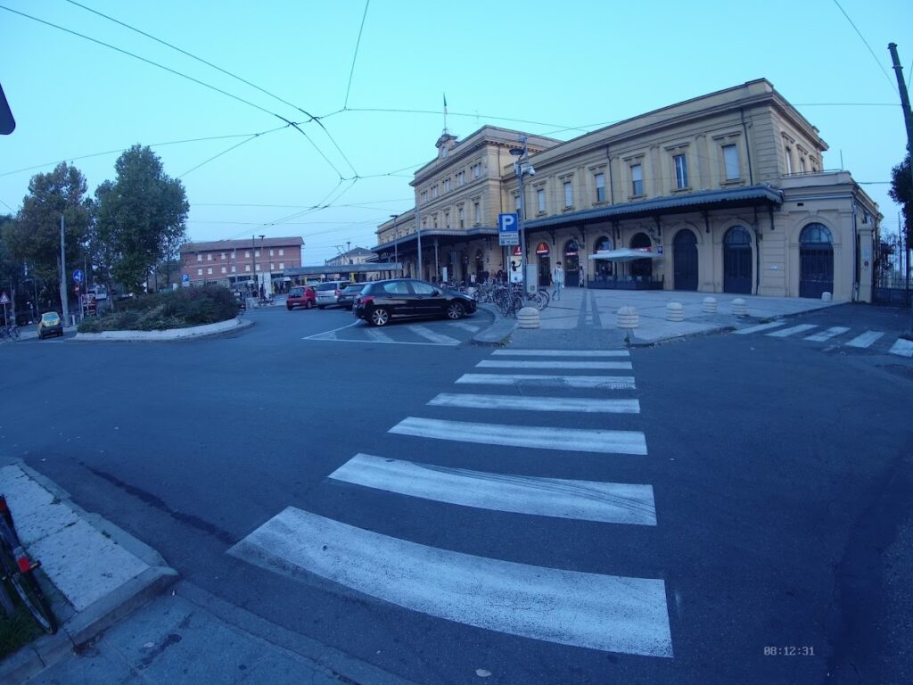 Modenna Historic train station facade with pedestrian crossing and parked vehicles, under a clear blue sky.