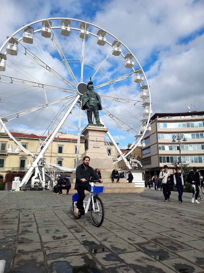 A man rides a bike near a statue and a Ferris wheel, capturing a vibrant urban scene on a cloudy day.