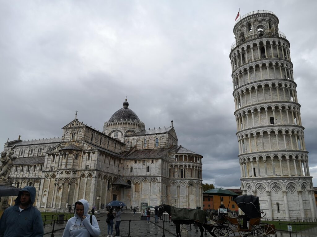 View of the Leaning Tower of Pisa alongside the Pisa Cathedral in a cloudy sky, showcasing iconic Italian architecture.
